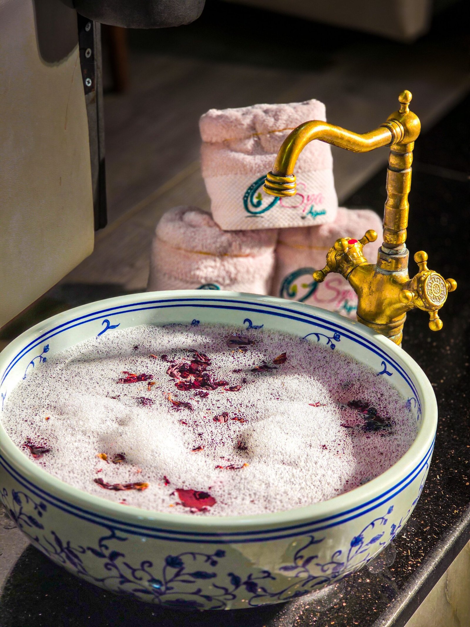 A luxurious spa bowl with rose petals and foam, accompanied by rolled towels and a vintage faucet.